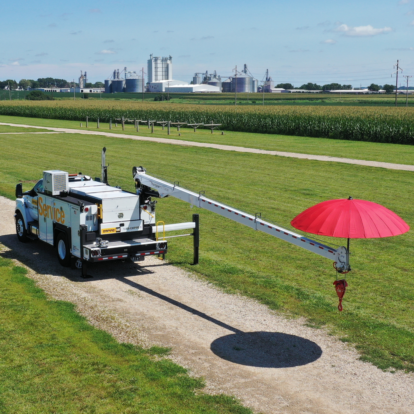 A white utility service truck with an extended boom arm parked on a dirt path in a grassy field, holding a large red umbrella. Cornfields and industrial silos are visible in the background under a blue sky.