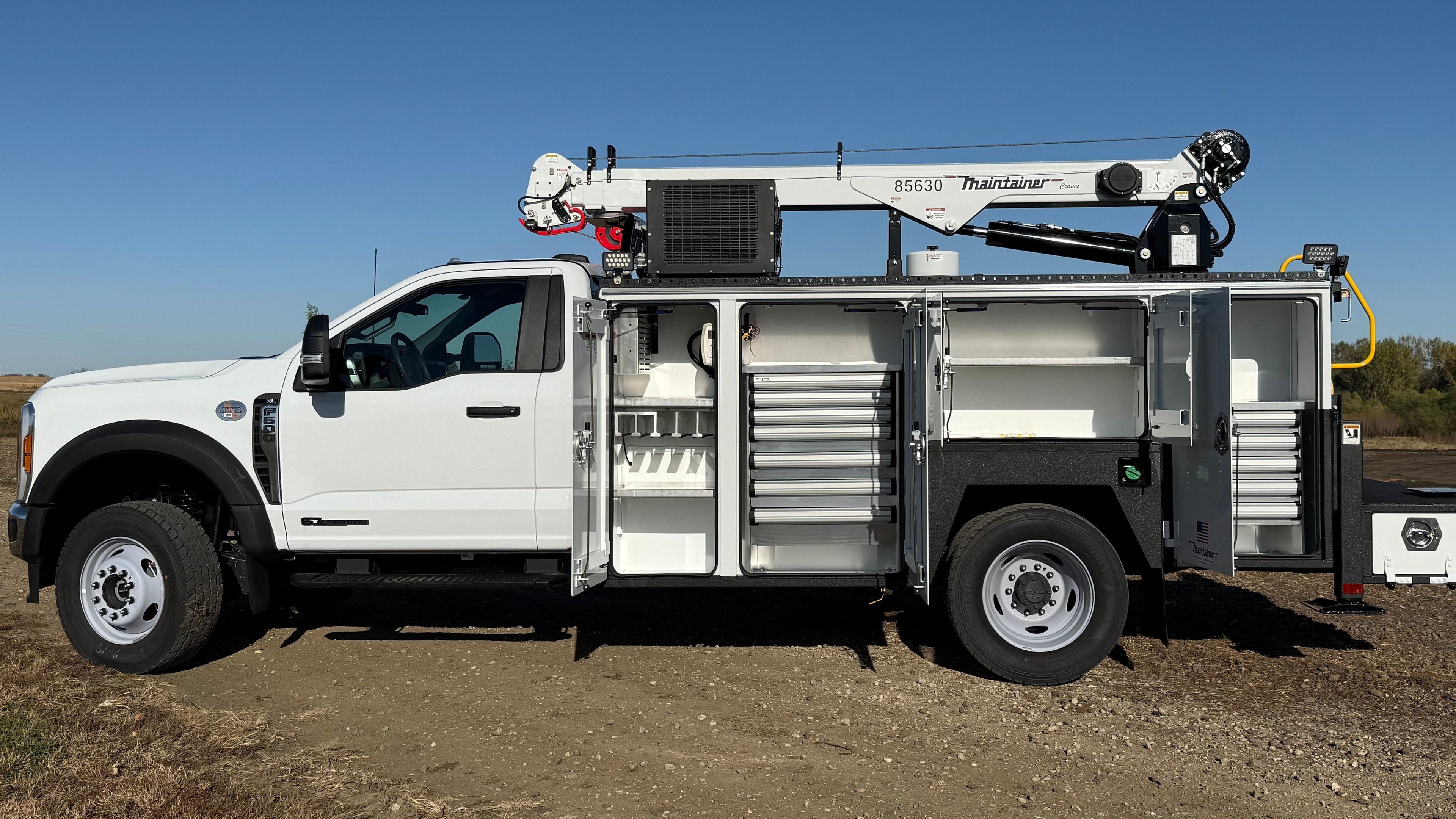 White utility truck with crane with organized truck compartments on a dirt road under a clear blue sky.